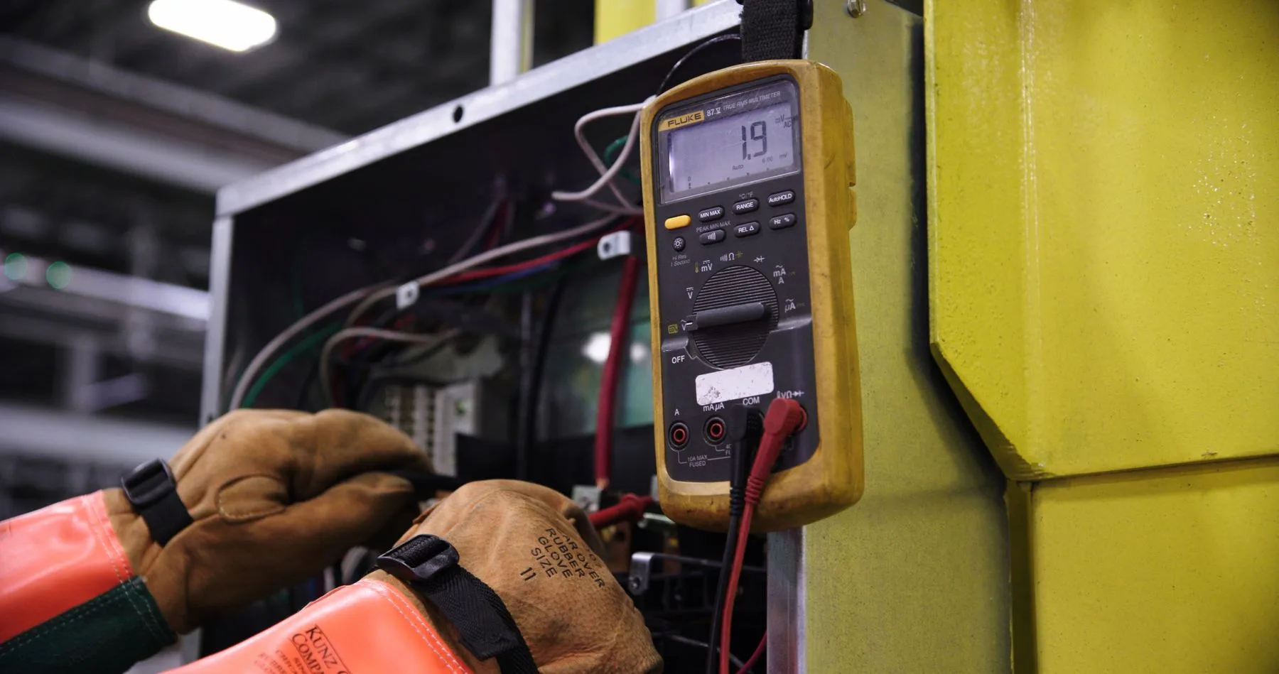 Gloved hands using a meter beside electrical controls