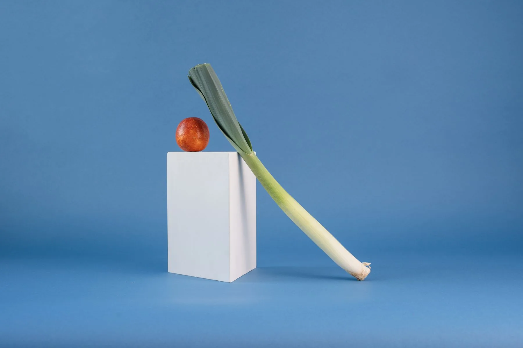 A leek and a piece of fruit arranged on a white plinth against a blue background.